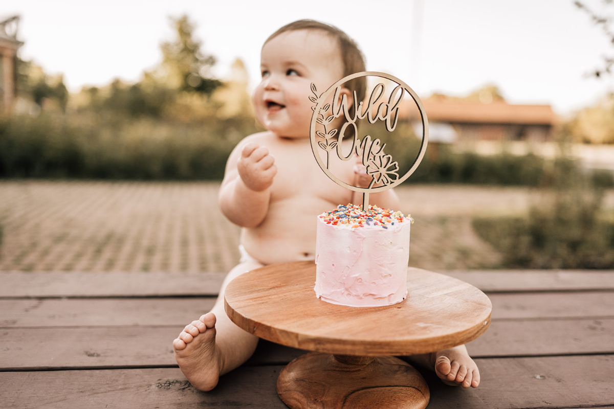 Baby sitting beside small cake on stand, smiling outdoors with soft golden light