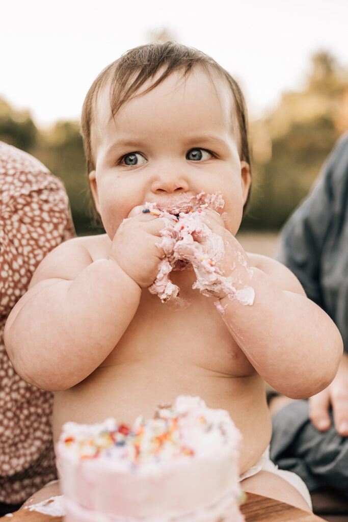 Baby grabbing handfuls of frosting, laughing while sitting with cake during outdoor session