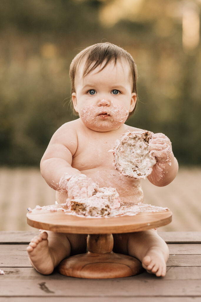 Baby covered in frosting holding cake, looking at camera during messy outdoor birthday moment