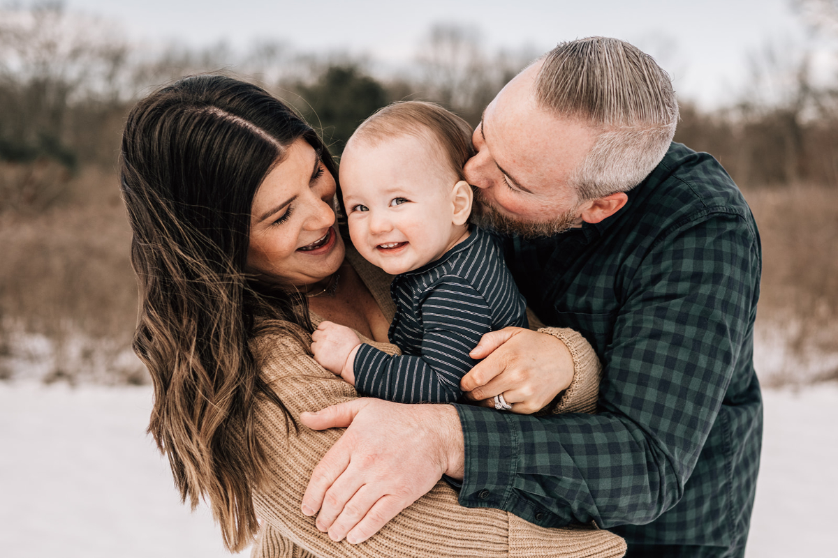 Parents hugging baby close outdoors in snow, smiling and sharing warm family moment together