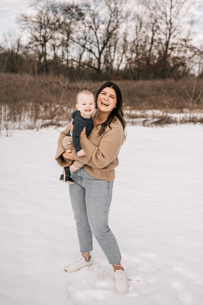 Mom holding baby while standing in snowy field, both smiling during relaxed outdoor session