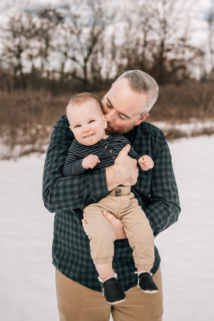 Dad holding baby and kissing cheek outdoors in winter, capturing playful and loving moment