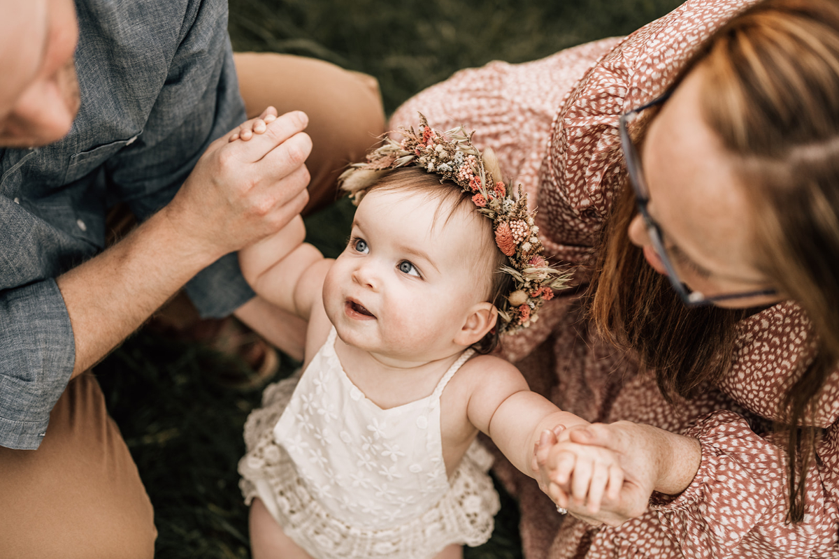 Baby looking up at parent while holding hands, wearing floral crown during warm outdoor family moment