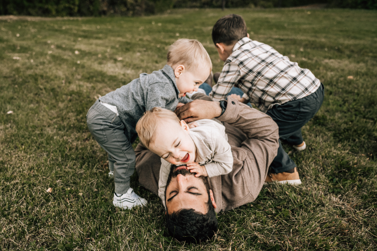 Father lying in the grass while his young children climb, laugh, and play around him outdoors.