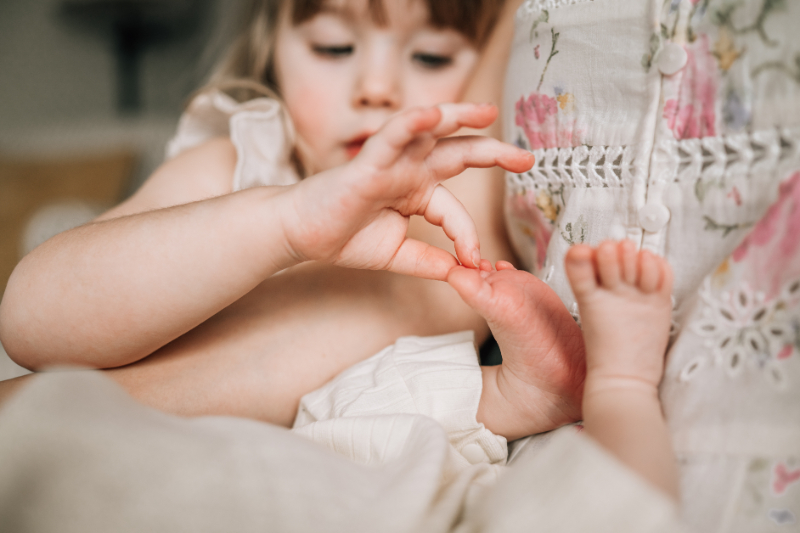 Toddler gently touching newborn’s tiny toes, showing curiosity, connection, and a quiet sibling moment.