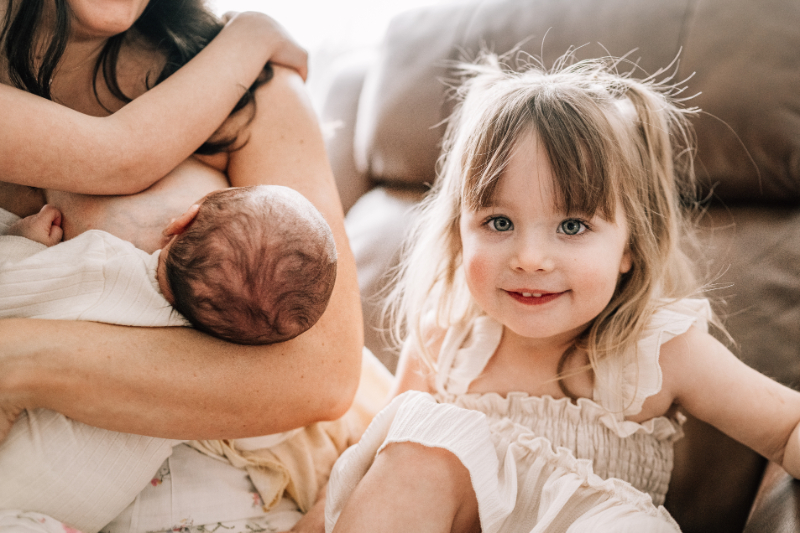 Mother holding newborn while sibling sits close during newborn photos at home, showing connection and curiosity.