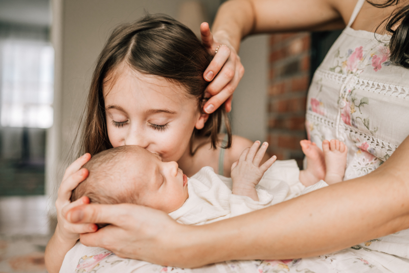 Sibling kissing newborn during newborn photos at home, capturing tender connection and quiet, loving moment.