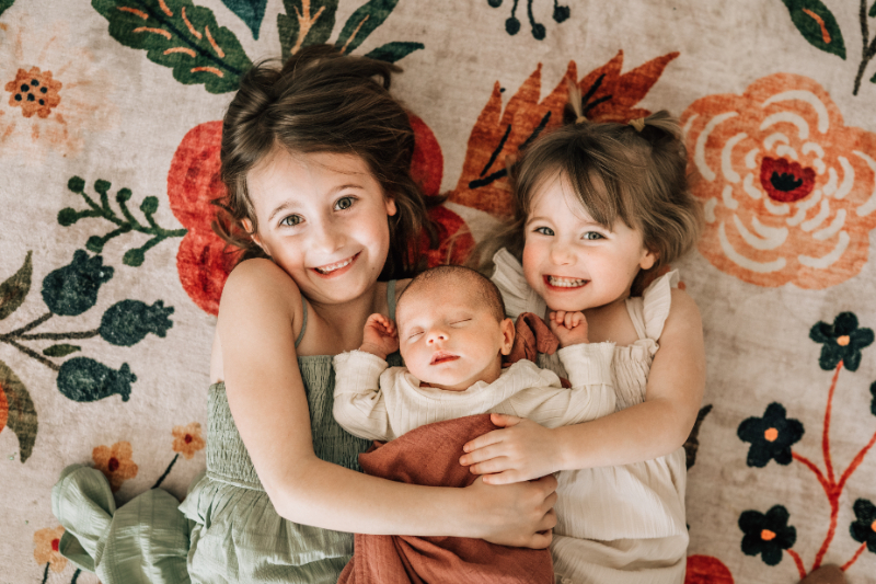 Siblings holding newborn on patterned blanket during newborn photos at home, capturing playful connection and warmth.