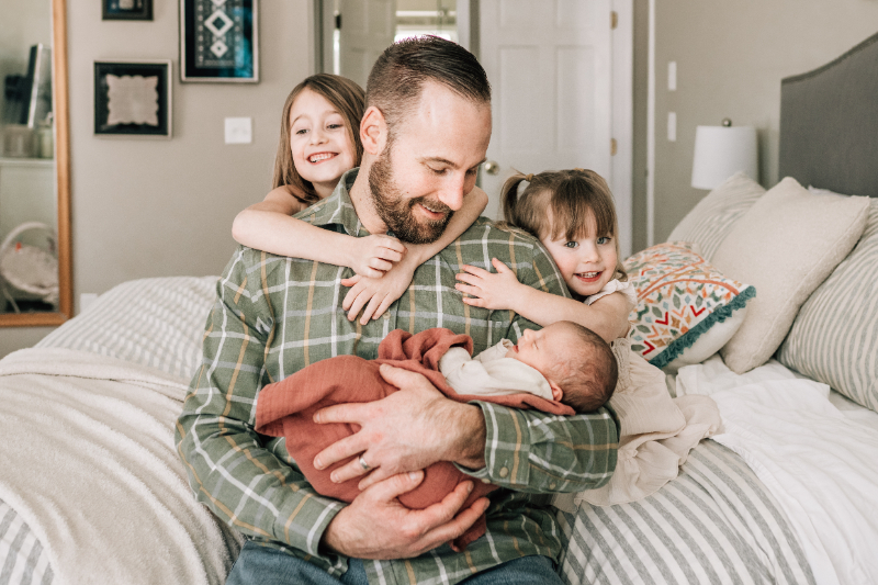 Father holding newborn with siblings on bed during newborn photos at home, showing joy and family connection.