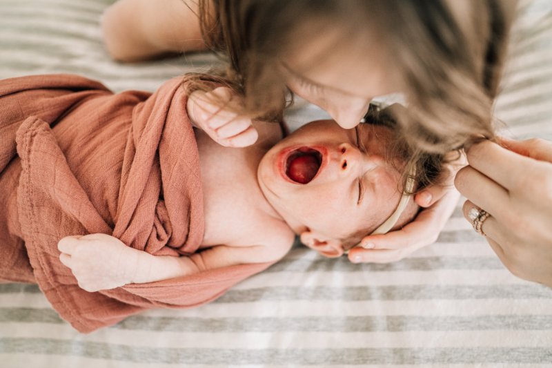 Sibling leaning in while newborn yawns, capturing a candid, emotional moment and real family connection.