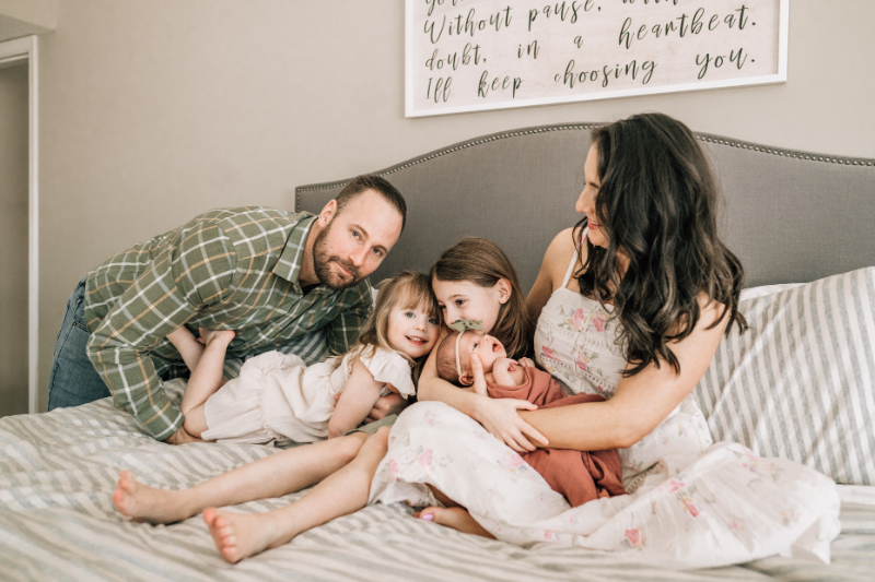 Family of four cuddling on bed during newborn photos at home, capturing relaxed, connected moments together.