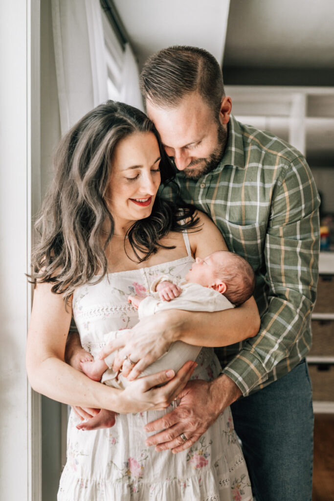 Mother and father holding newborn by window during newborn photos at home, capturing soft, intimate connection.