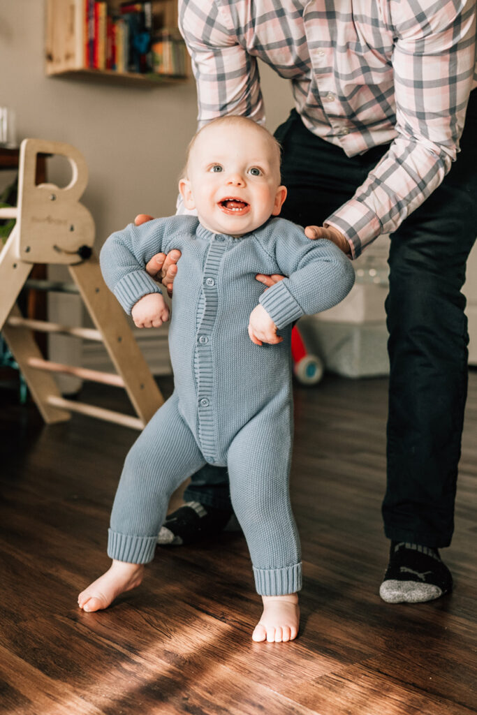 First birthday family photos of a one-year-old taking supported steps during a relaxed in-home birthday session.