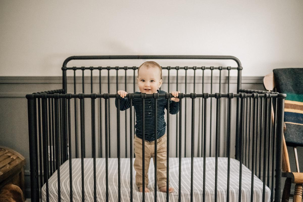 One-year-old standing in a crib while smiling during a relaxed in-home lifestyle photography session.
