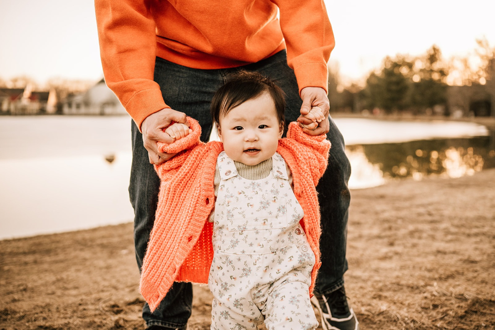 First birthday family photos of a toddler taking first steps while holding a parent's hands outdoors.
