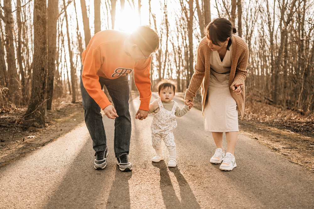 First birthday family photos of parents walking hand in hand with their one-year-old on a wooded trail.
