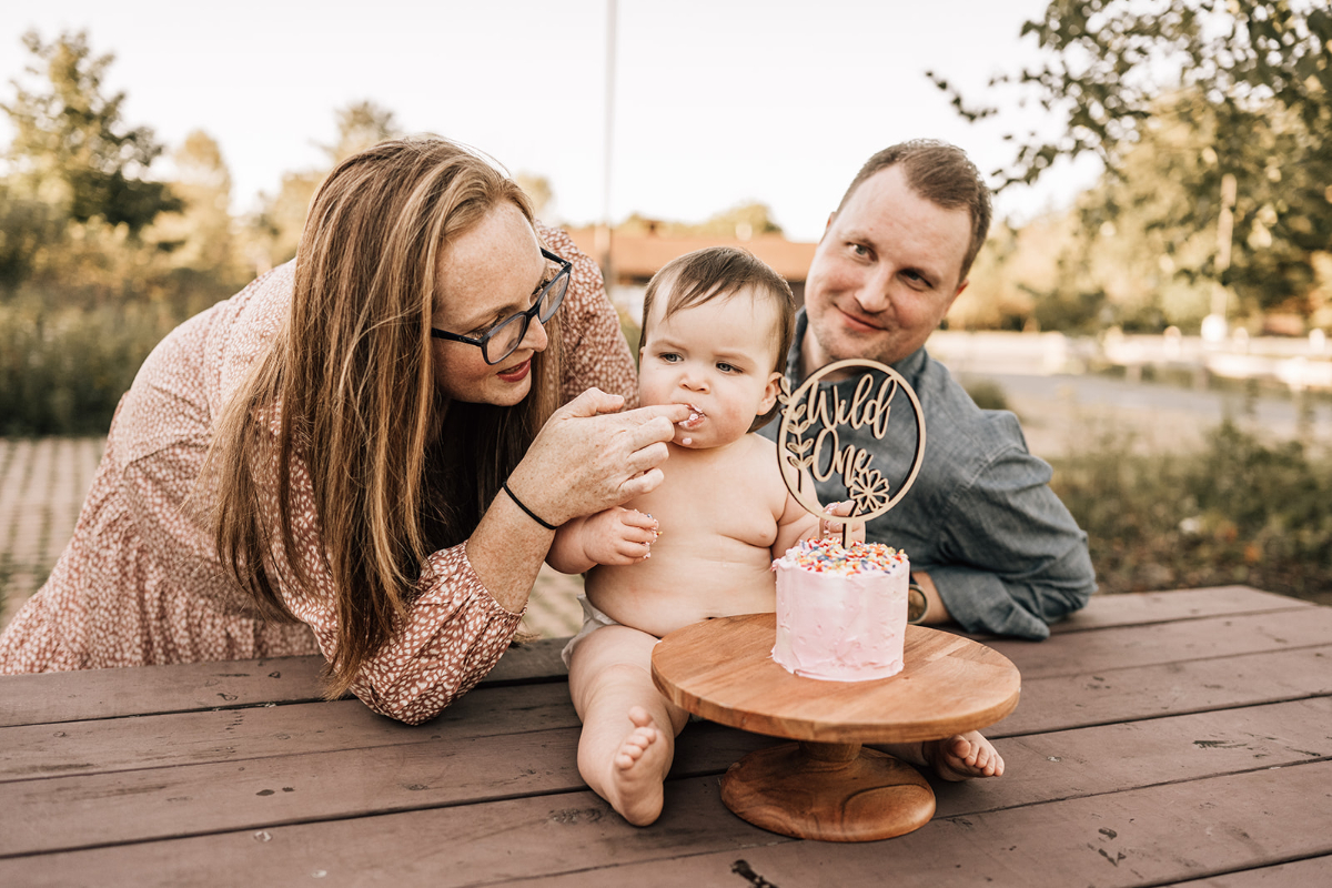 First birthday family photos of a baby enjoying a small cake while parents smile beside them outdoors.