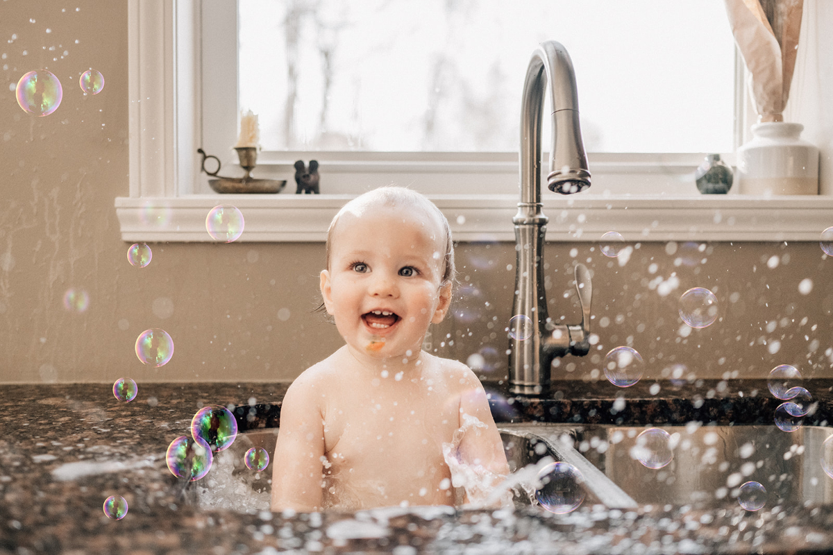 One-year-old splashing in a kitchen sink bath while playing with bubbles during an in-home birthday session.