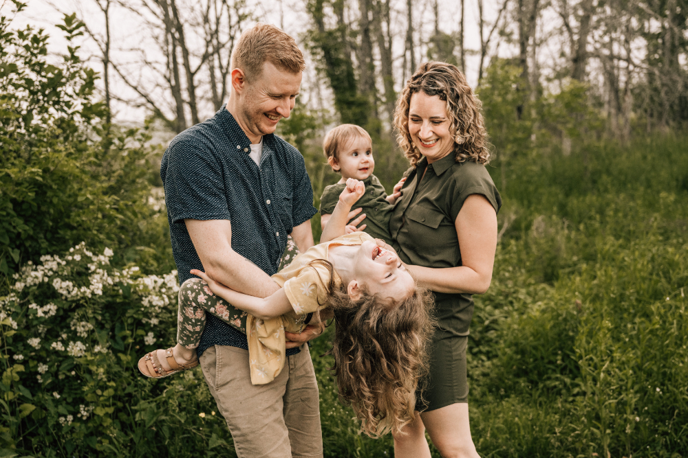 A family of four laughing together as a father playfully holds his daughter upside down outdoors.