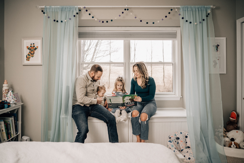 Parents and their two children sitting by a bedroom window reading a book together.