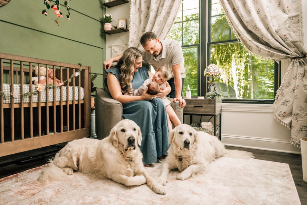 Parents cuddle their newborn and young child in the nursery while their two dogs rest nearby.