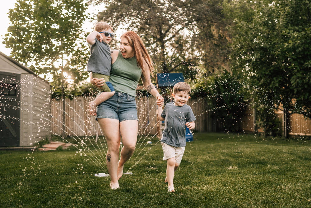 Outdoor family photo ideas with a mom and her children running through backyard sprinklers on a summer evening.