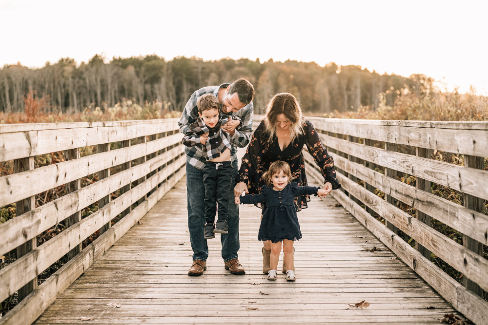 Family walks hand in hand across a wooden bridge during a playful golden hour session.