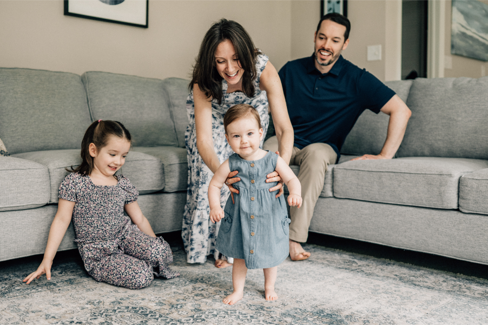 Mother helping her baby stand while the rest of the family gathers together in the living room.