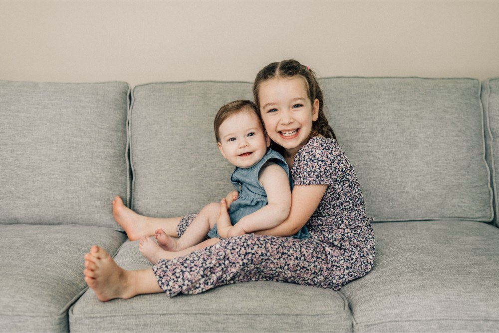 Two young sisters smiling and cuddling together on the living room couch.