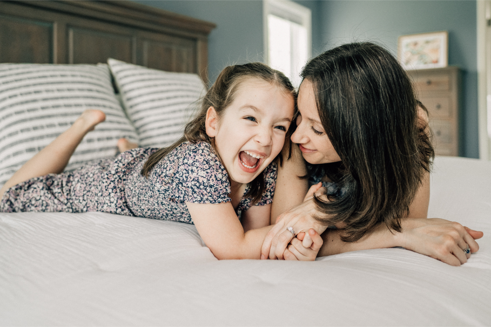 At home family photos of a mother and daughter laughing together while cuddling on the bed.