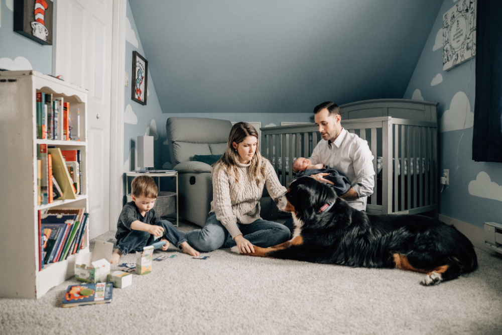 At home family photos of parents relaxing with their toddler, newborn, and dog in the nursery.